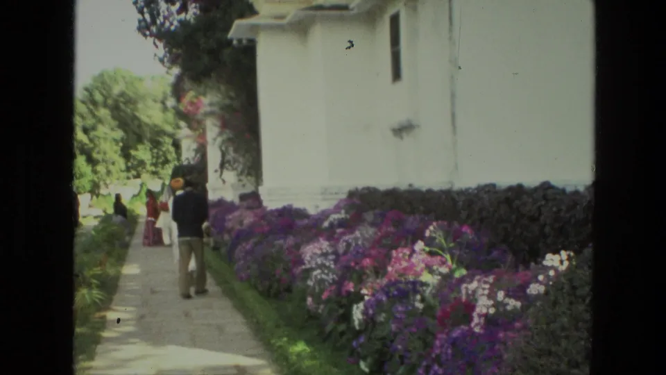 1974 India home-movie frame showing tourists on a stone path beside purple flower beds and a white building.