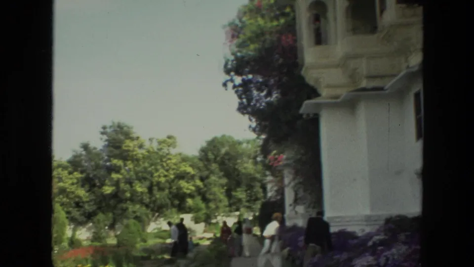 1974 India frame showing tourists from behind on a garden path beside a white building and colorful plantings.