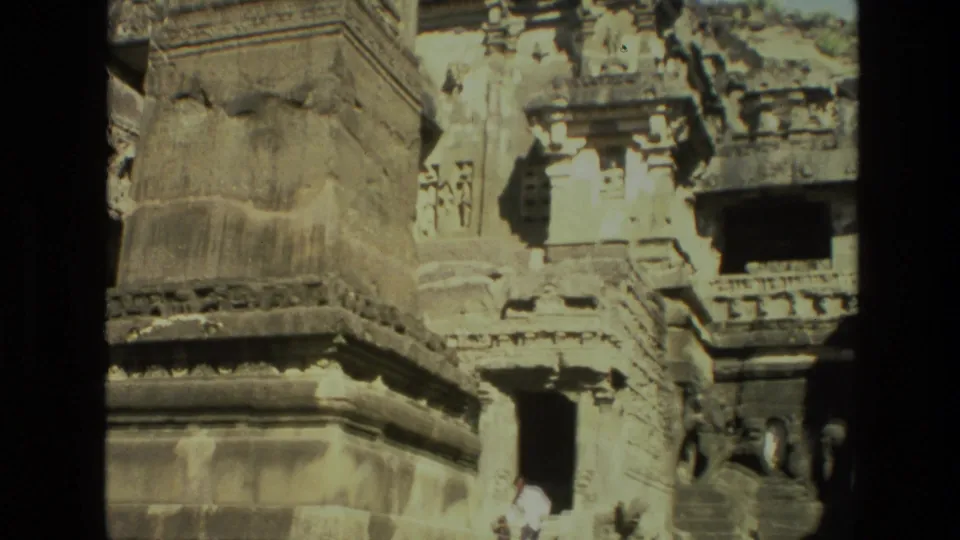 1974 India 8mm frame showing carved temple stonework, a dark entrance, steps, and a small visitor in light clothing near the doorway.