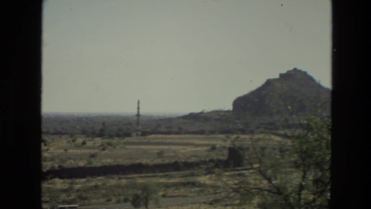 Dry plain seen through dark vehicle window edges with a distant tower and rocky hill.