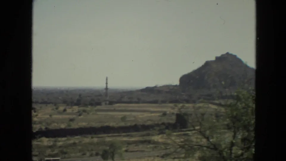 1974 India 8mm frame showing an arid plain from a vehicle window, with a distant tower, sparse trees, and a rocky hill.