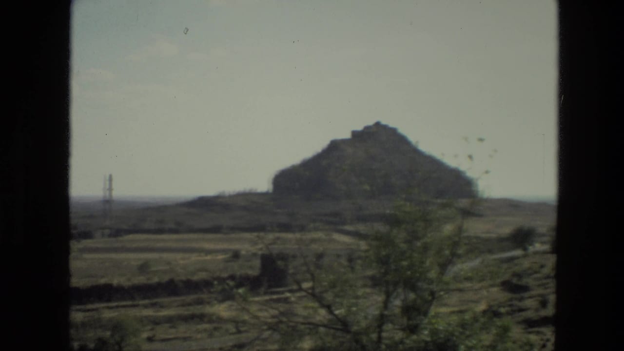 Rocky hill and dry fields framed by a dark window edge, with a distant tower at left.