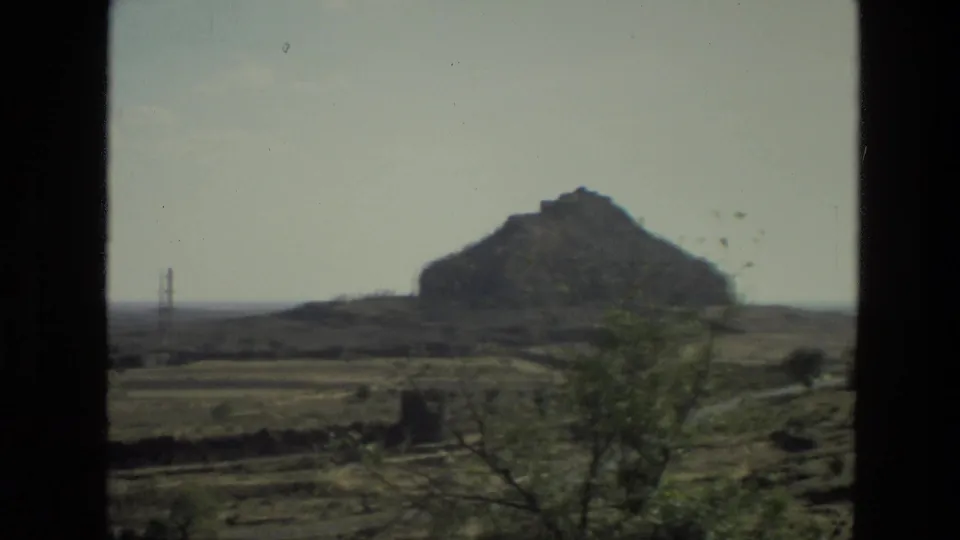1974 India travel frame with a rocky hill, dry fields, sparse vegetation, and a distant tower viewed through a vehicle window.