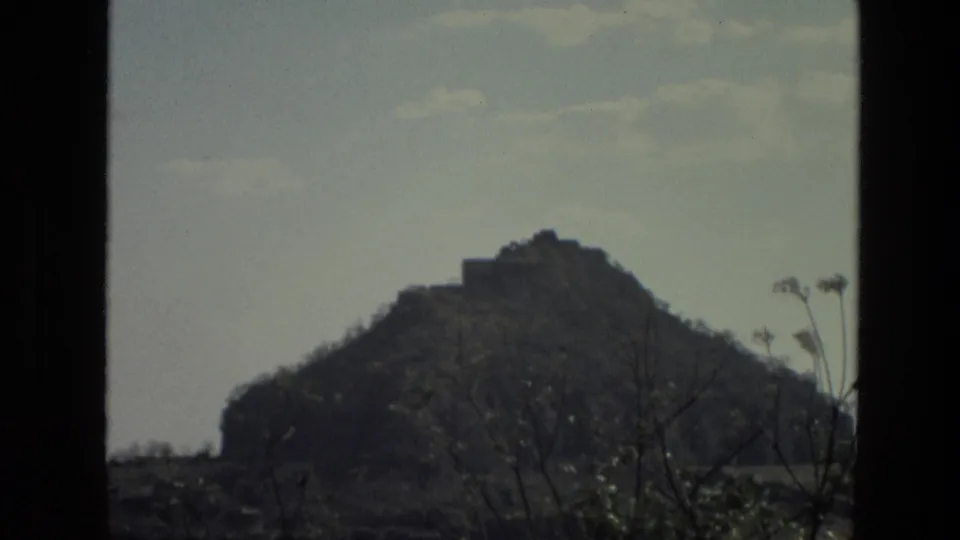 1974 India frame showing a rocky hill and hilltop structure beyond foreground branches, with a pale sky and curving road nearby.