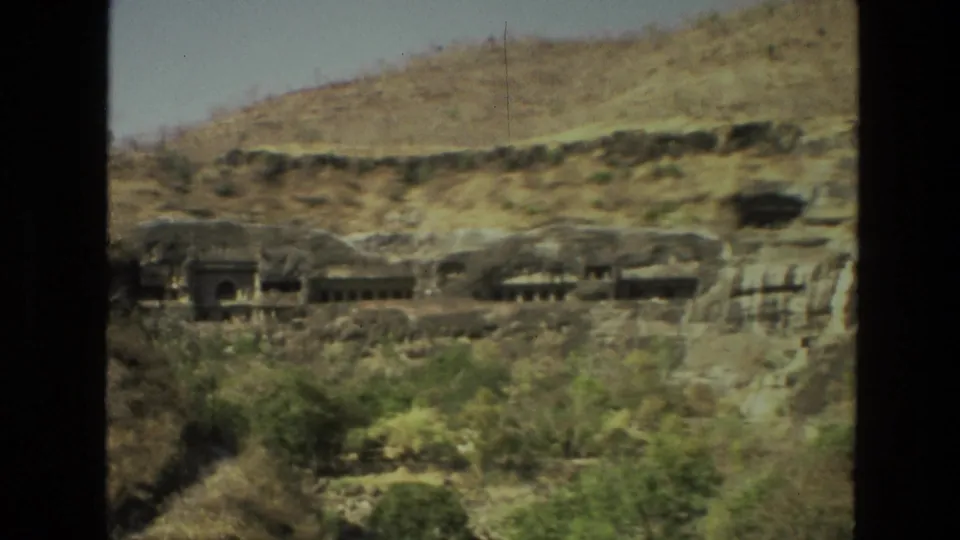 1974 8mm view in India showing rock-cut cave temple openings across a dry hillside, framed by dark film borders.
