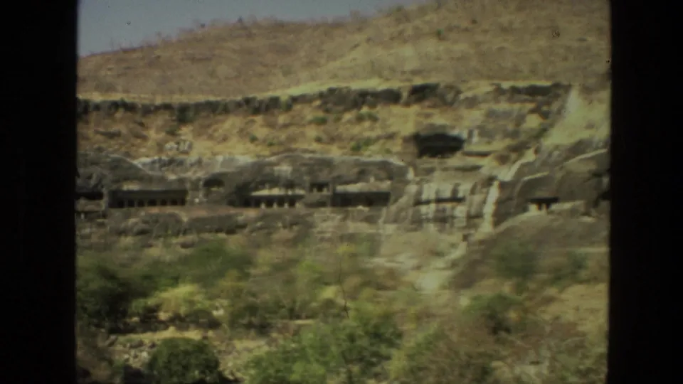 A distant 8mm frame of Indian cave temple architecture, with carved entrances, shadowed openings and scrub vegetation below.