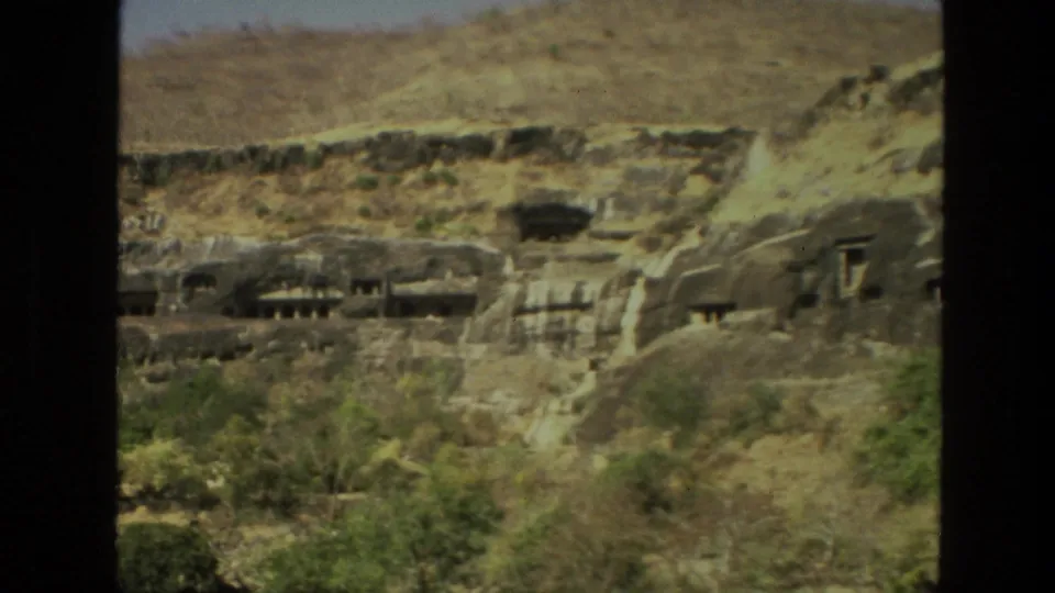 1974 home-movie frame in India showing rock-cut cave temples set into brown stone above trees and brush.