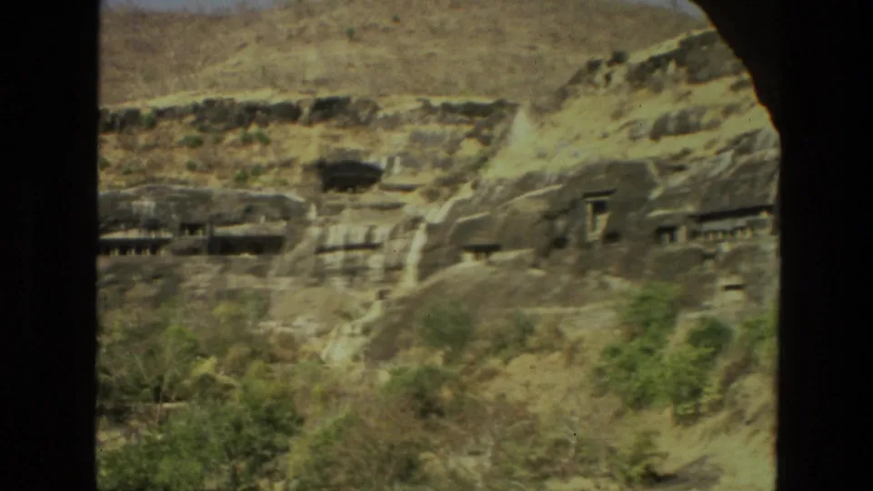 Distant 8mm view of Indian cave temples in a rugged hillside, with dark openings, carved stone surfaces and green foreground growth.