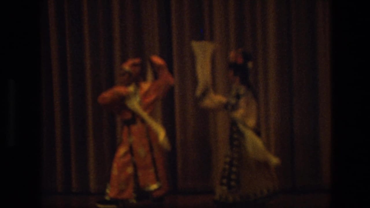 Two costumed dancers perform with white scarves on a dim stage before a brown curtain.