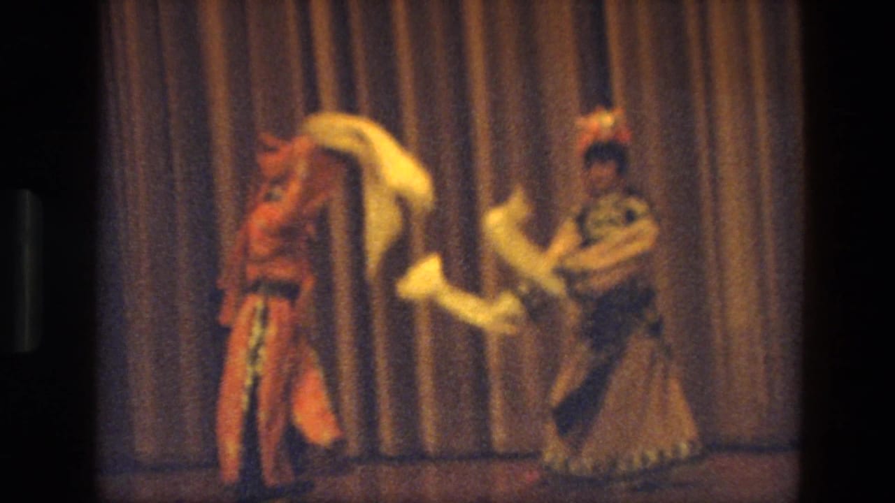 Two dancers in ornate costumes face each other while holding long white scarves on stage.