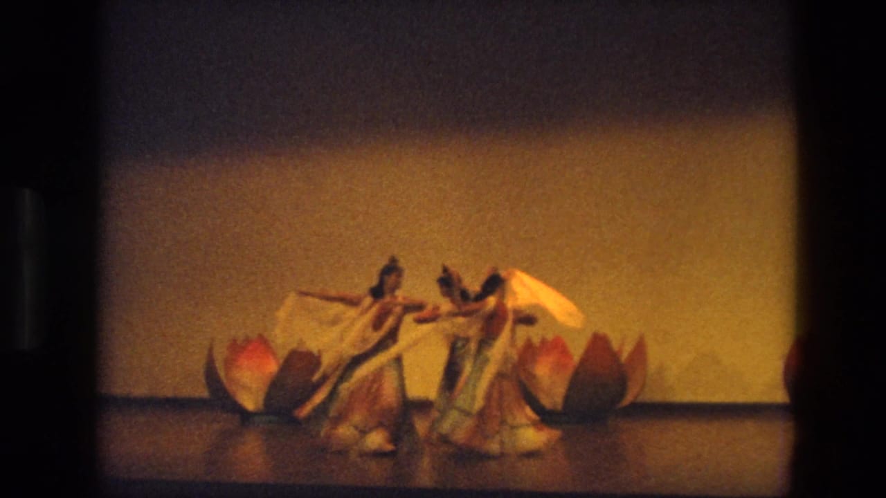Three women perform a staged dance in India in 1974, wearing flowing costumes near red and gold lotus-shaped props.
