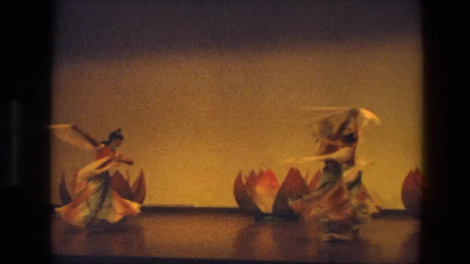 Costumed dancers continue a 1974 India stage routine, with one dancer turning near large lotus props.