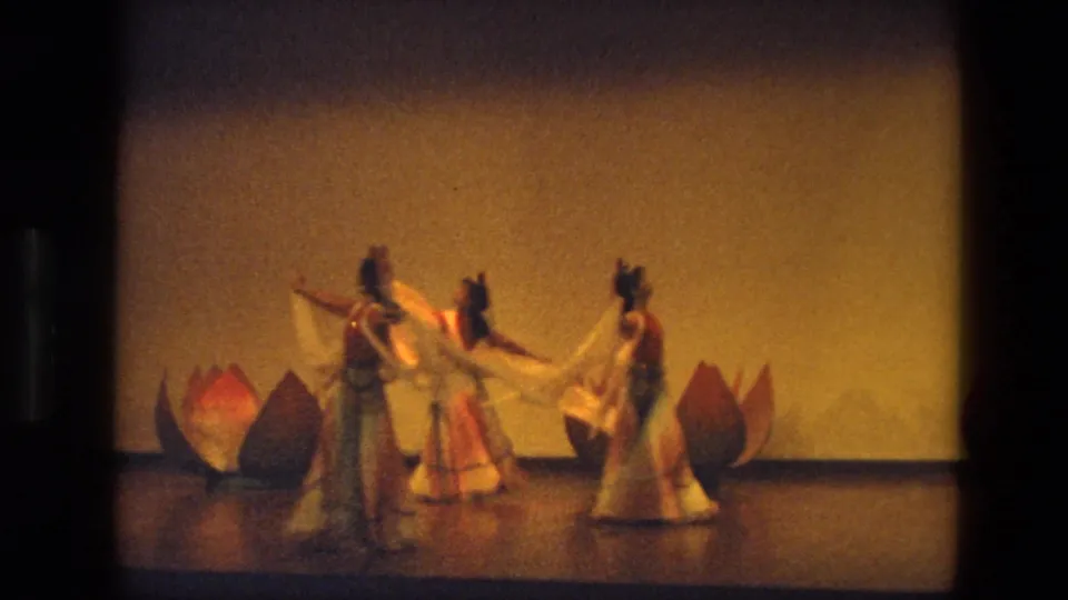 Three women in traditional-style costumes gather mid-routine on a 1974 India stage with lotus props behind them.