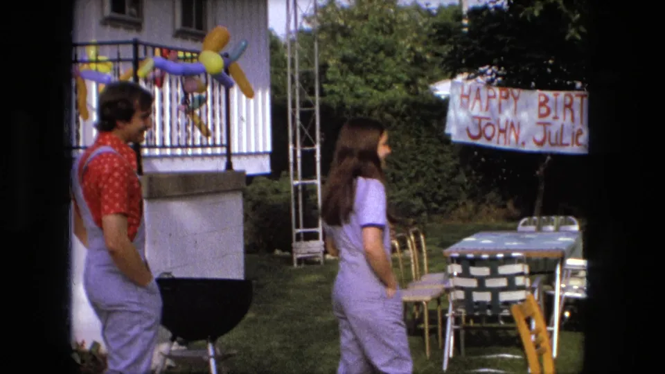 A man in a red shirt and overalls stands with a woman near a grill and handmade birthday banner in a 1976 Fort Wayne backyard.