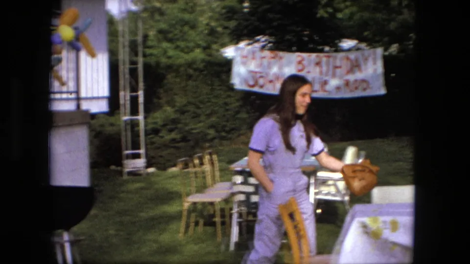 A woman in a purple jumpsuit carries items beside a table, chairs, and handmade birthday banner in Fort Wayne, Indiana, 1976.