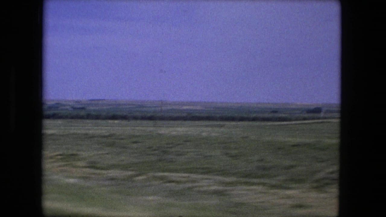 Green fields and a distant horizon seen through dark film or window edges.