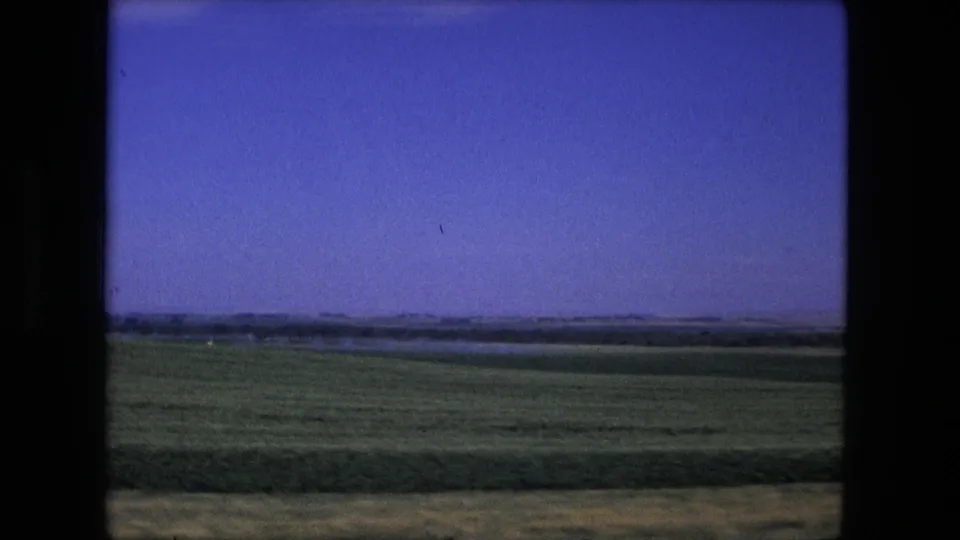 Fort Wayne, Indiana area landscape in 1976, showing open green farmland, a distant tree line, and clear blue sky from a moving vehicle.