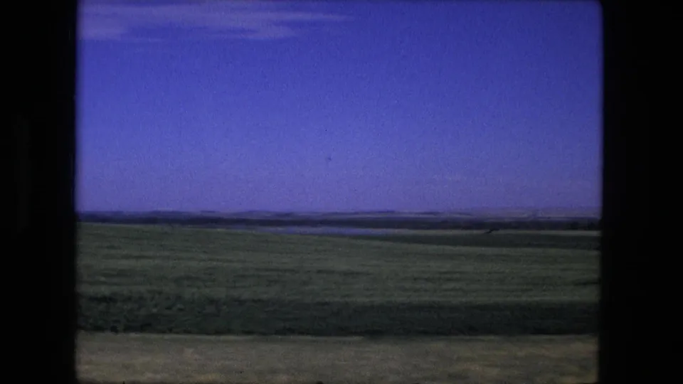 A 1976 home-movie view of flat green farmland near Fort Wayne, with a low horizon and dark film framing visible at the edge.