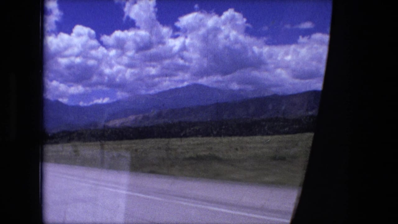 Rural highway seen through a car window with fields, hills, clouds, and a dark windshield pillar.