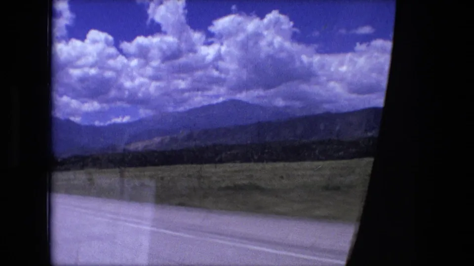 1976 8mm home-movie frame from a moving vehicle, showing open rural roadside, distant hills, bright clouds, and part of the car interior.