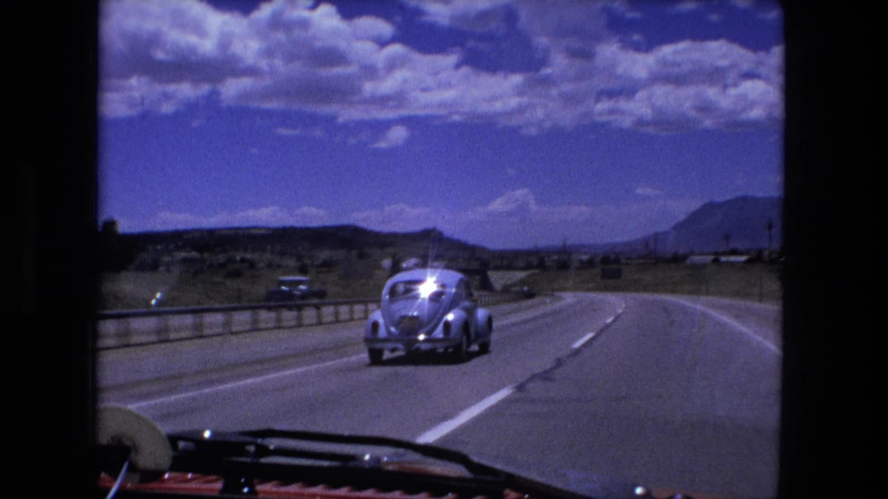 1976 8mm road-trip frame showing a light blue Volkswagen Beetle on a broad rural highway, filmed from another moving car.