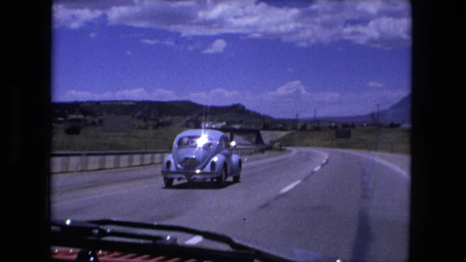 1976 8mm frame with a light blue Volkswagen Beetle continuing along a curving highway, with open land and low hills beyond.
