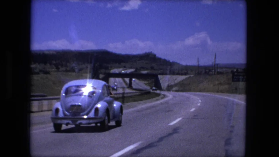 1976 8mm home-movie frame showing a light blue Beetle near a highway overpass, with dry roadside terrain and distant hills.