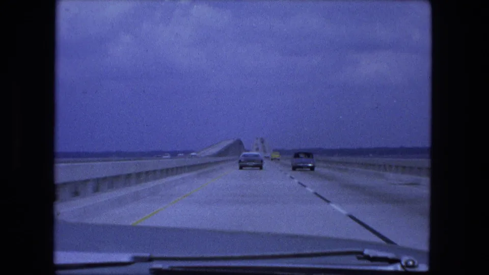 1974 8mm driver-perspective view across a long bridge over water, with cars ahead under a blue-tinted sky.