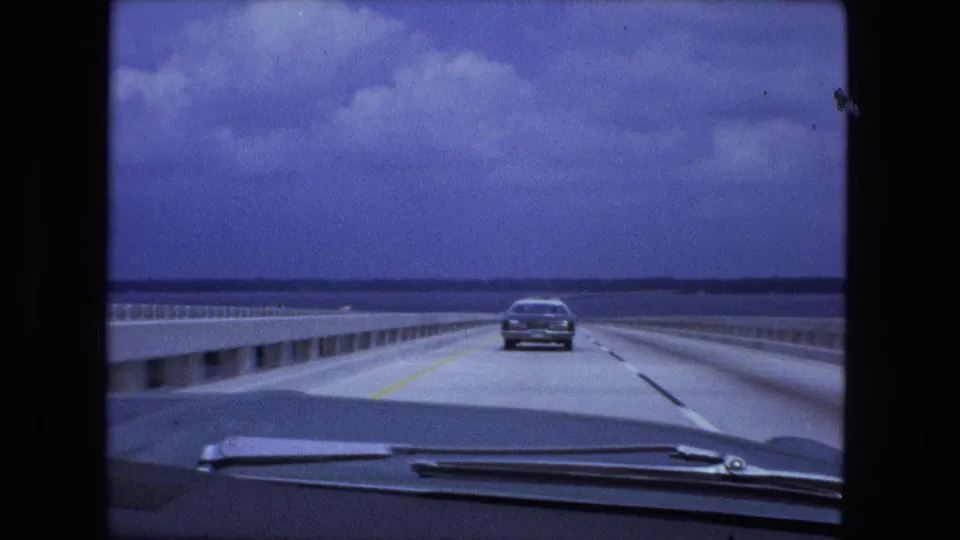 Driver-view 8mm frame of a car ahead on a long bridge crossing, with water and low shoreline in the distance.