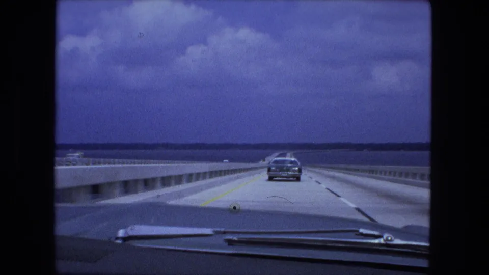 1974 8mm road-travel view from inside a car, following another vehicle across a bridge over open water.