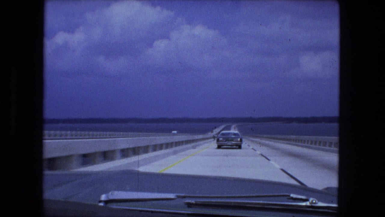 Forward view from a car crossing a long bridge with a sedan ahead and water beside the roadway.