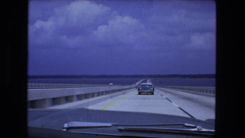 1974 home-movie view from inside a car traveling over a long bridge, with a sedan ahead, guardrails, water, and a blue sky.
