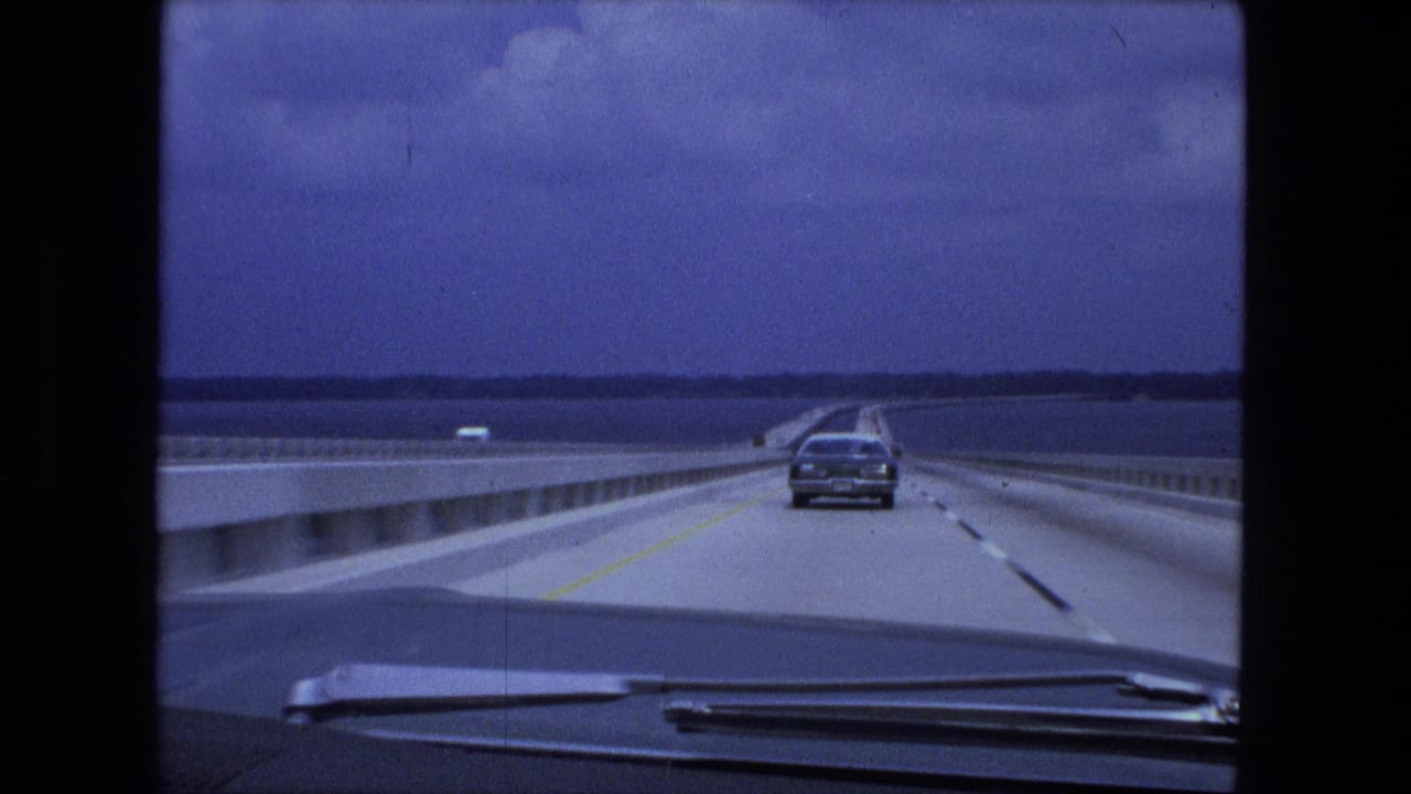 Forward 8mm view across a bridge in the Fort Wayne-labeled 1974 clip, showing a sedan ahead, water, guardrails, and distant land.