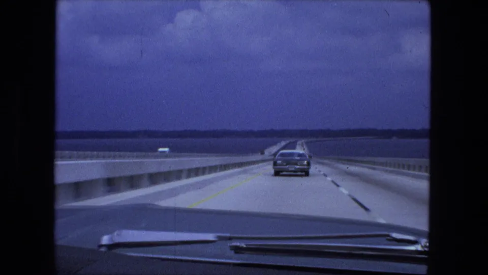 Forward 8mm view across a bridge in the Fort Wayne-labeled 1974 clip, showing a sedan ahead, water, guardrails, and distant land.