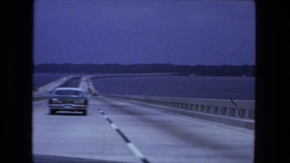 The following car nears a sedan on the bridge, with water, guardrails, lane stripes, and a distant tree line visible in the 8mm frame.