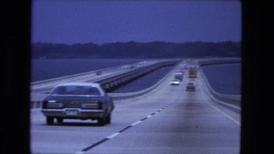 A 1974 8mm bridge-driving view shows a sedan ahead, a gentle road curve, other distant cars, guardrails, water, and shoreline.