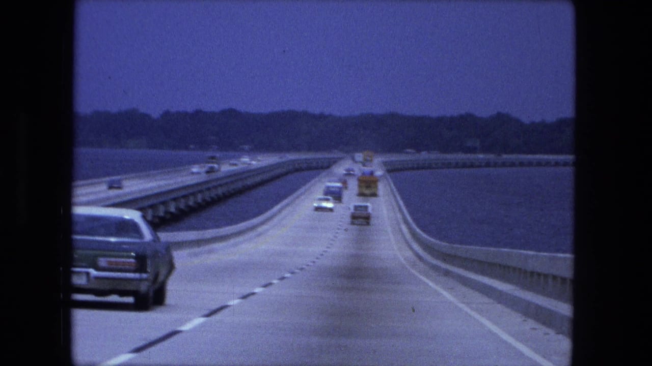 Cars travel on a bridge or causeway over water with concrete barriers and a distant shoreline.