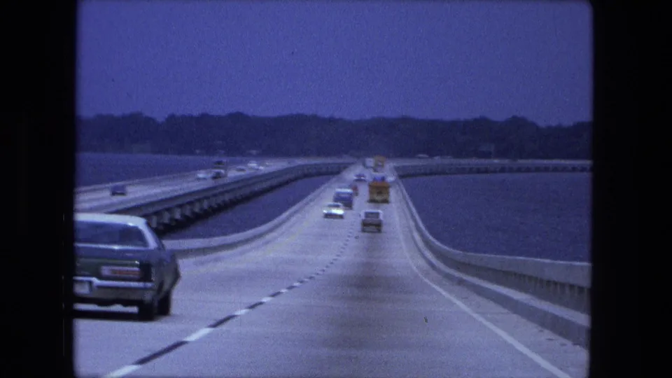 1974 home-movie view from a moving car on a bridge or causeway over water near Fort Wayne, Indiana, with traffic ahead and a tree-lined shore.
