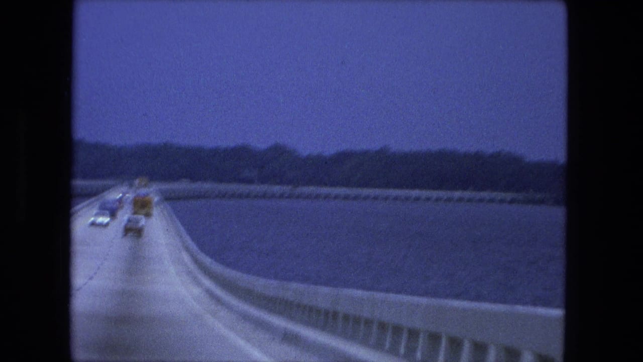Moving-vehicle view of traffic receding along a 1974 bridge or causeway, with water, concrete guardrail, and dark tree line in the distance.