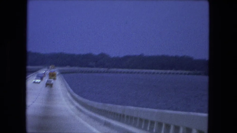 Moving-vehicle view of traffic receding along a 1974 bridge or causeway, with water, concrete guardrail, and dark tree line in the distance.