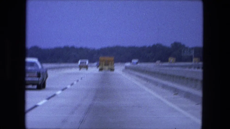 1974 8mm travel shot from inside a moving vehicle, following cars and a truck along a bridge or causeway with low railings and distant trees.