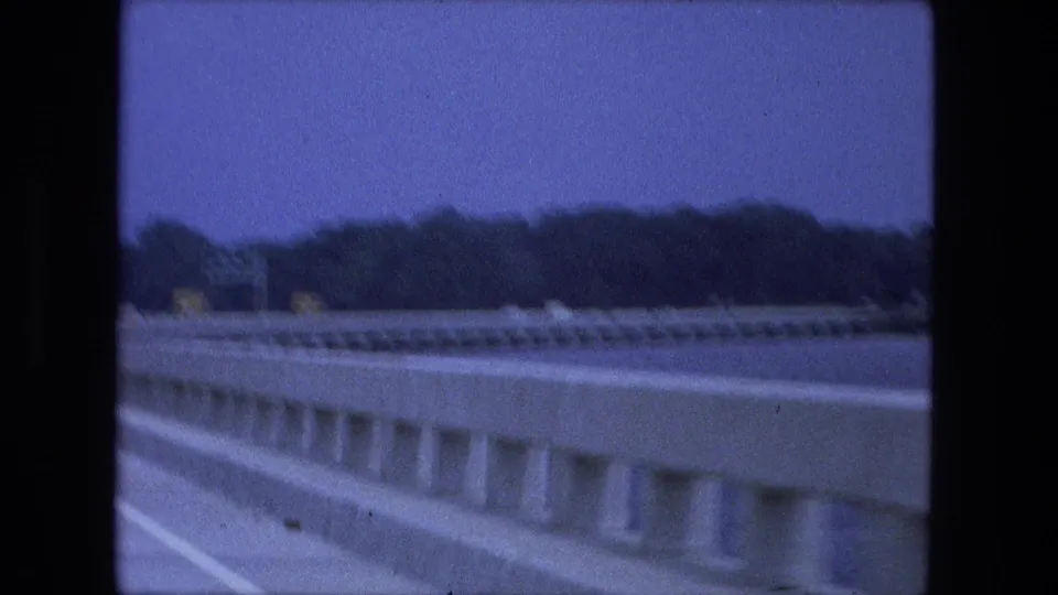 End-frame view from the moving car emphasizes the concrete bridge barrier, water beside the roadway, and wooded shoreline under a blue sky.