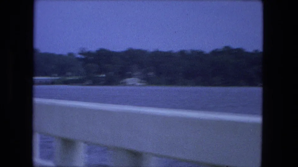 1974 Fort Wayne home-movie frame showing blue water, distant trees, and a pale barrier from a moving vehicle viewpoint.