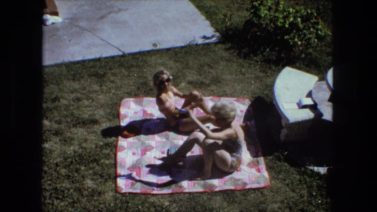 Two women sit on a patterned blanket on grass beside white patio furniture.