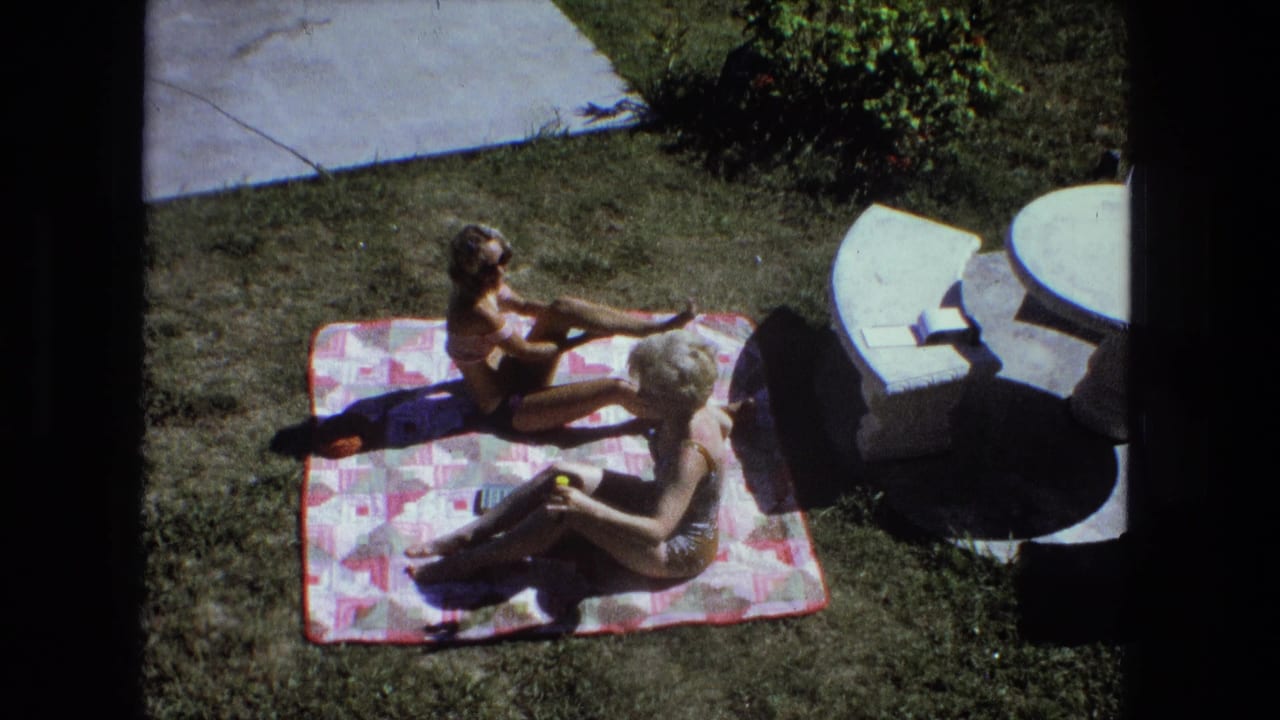 Two women in swimsuits sit on a patterned backyard blanket in 1974 Fort Wayne home-movie footage.