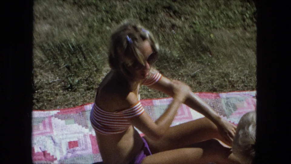 Close 8mm view of a woman in sunglasses applying sunscreen on a backyard blanket in Fort Wayne, Indiana, 1974.
