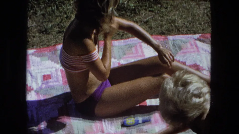 1974 Fort Wayne home-movie frame showing two women on a backyard blanket, with one rubbing sunscreen on her arm.