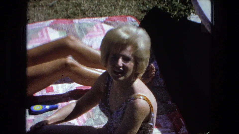 A blonde woman smiles toward the camera from a patchwork blanket during the 1974 Fort Wayne sunbathing home movie.