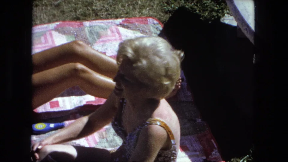 1974 Fort Wayne home-movie still of a woman in profile on a backyard blanket, with another sunbather partly visible behind her.