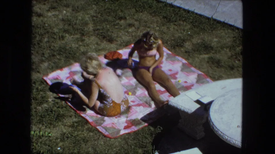 Wide 1974 Fort Wayne backyard view of two women in swimsuits seated on a patterned blanket beside grass and a round patio table edge.